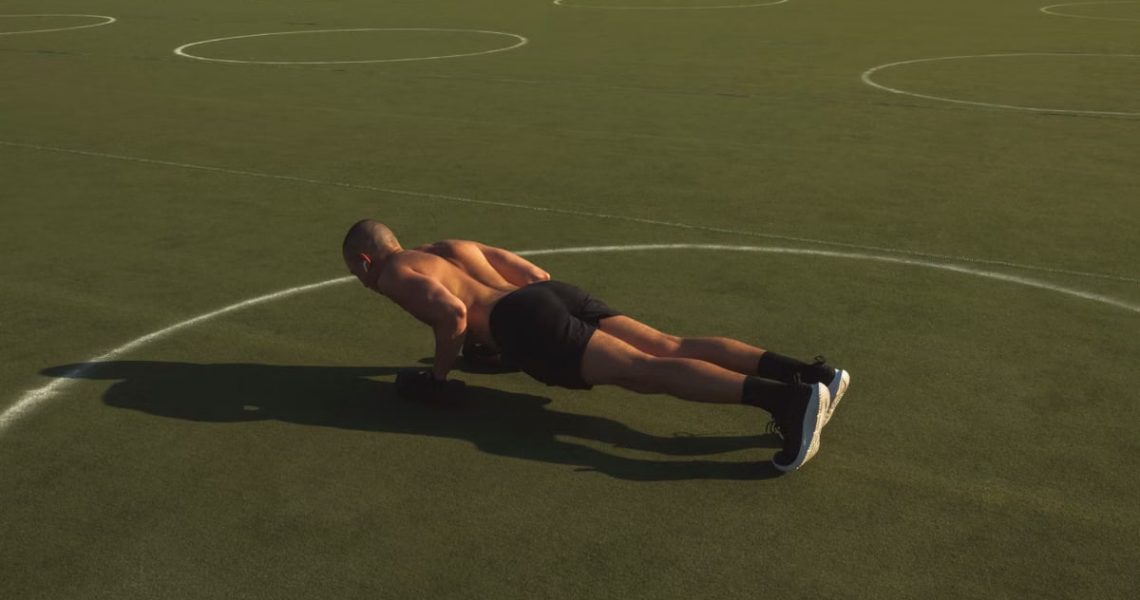 A shirtless man performing push-ups on a green field under bright daylight.